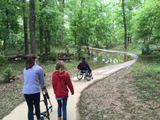 3 people, including one using a walker and one chair user, walk on a trail that moves into the woods. Part of the trail is under water.